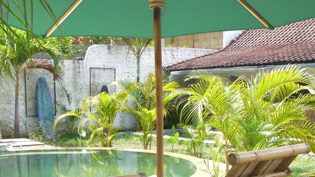 A pool area with two bamboo lounge chairs, a large green umbrella, and a stone wall in the background. - Palmhouse Boutique Hotel (Kuta)
