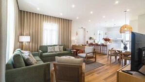 Een moderne woonkamer met een groene bank, een houten stoel en een televisie. Achtergrond is een keuken zichtbaar.