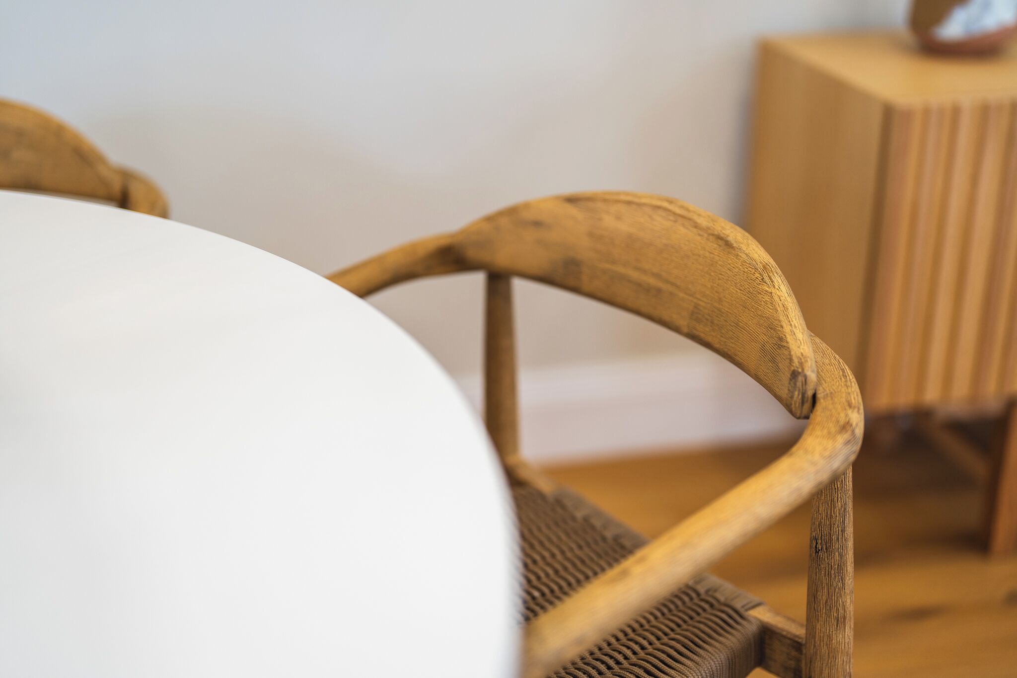 Primer plano de una silla de madera con asiento y respaldo trenzados, ubicada junto a una mesa blanca.