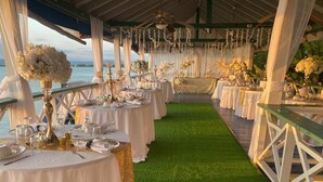 Un área de comedor al aire libre techada con mesas dispuestas para un evento formal, con vista a un cuerpo de agua.