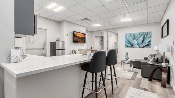 A modern kitchen with a marble countertop, black bar stools, and a dining area with a sofa and a lamp. - Stylish & Prestigious! 2 Bed/2 Bath-On The Avenue (Fountain Hills)