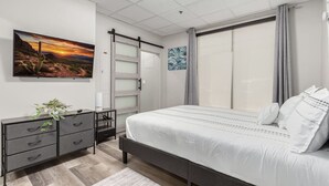 A bedroom with a bed, a dresser, a plant, a painting, and a window with curtains. - Stylish & Prestigious! 2 Bed/2 Bath-On The Avenue (Fountain Hills)