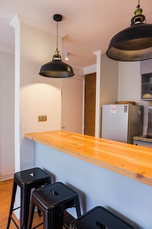 A kitchen with a wooden countertop, two black stools, a refrigerator, and pendant lighting.