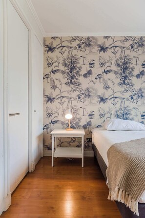 A bedroom with a bed, a bedside table with a lamp, and a floral wallpaper.
