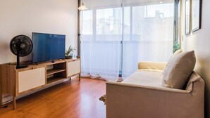 Una sala de estar con televisión, ventilador, sofá y ventana con cortinas de gasa.