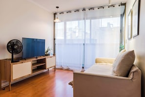 Una sala de estar con televisión, ventilador, sofá y ventana con cortinas de gasa.