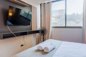 A hotel room with a flat-screen TV mounted on the wall, a standing fan, a neatly made bed with white linens, and a window with a view of buildings.
