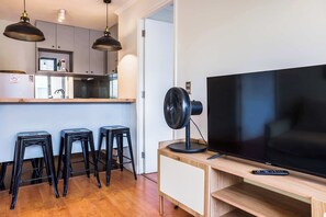 A modern kitchen with a microwave, a refrigerator, and a sink. There are three black stools and a wooden bar counter with a TV and a fan.