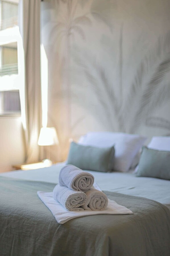 A neatly made bed with rolled towels and a white pillow, a lamp on a bedside table, and a window with curtains.