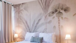 Un lit bien fait, avec une couette blanche et des oreillers, une table de chevet en bois avec une lampe, et un mur orné d’une fresque représentant un palmier.