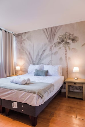 A neatly made bed with a white comforter and pillows, a wooden nightstand with a lamp, and a wall with a palm tree mural.