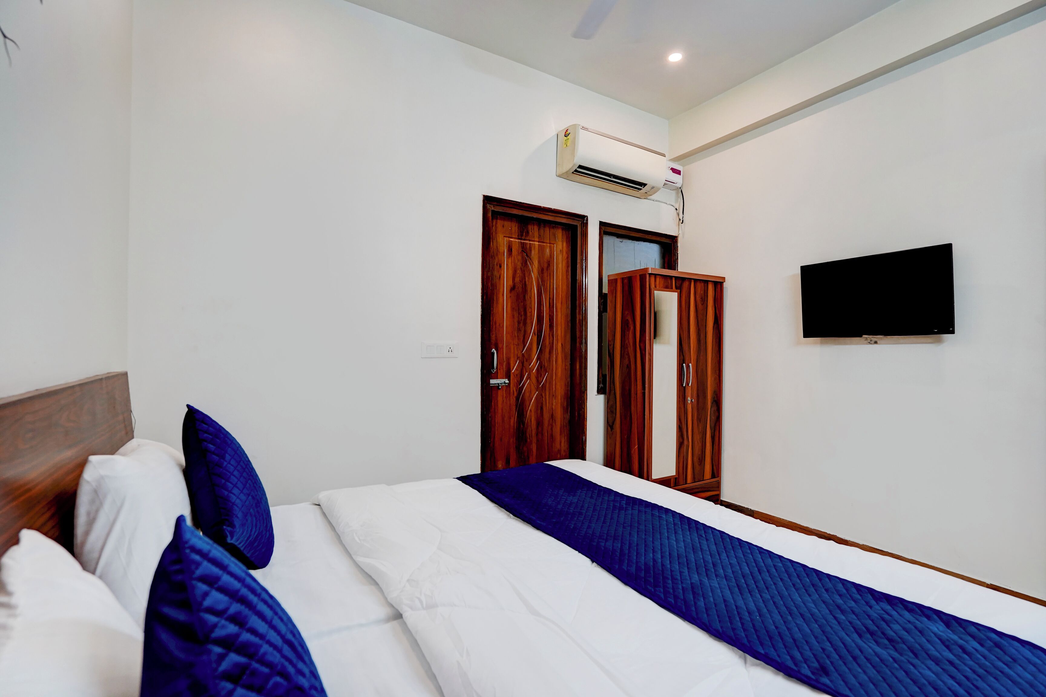 A hotel room with a bed, a TV mounted on the wall, and an air conditioning unit above the door.