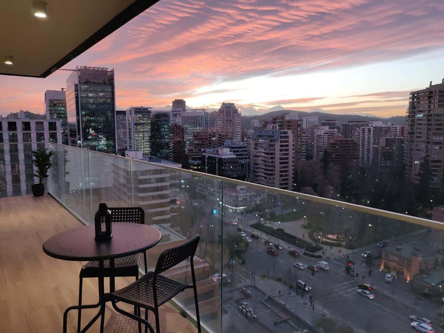 Un balcón con vista a la ciudad al atardecer, con edificios modernos y barandilla de vidrio.