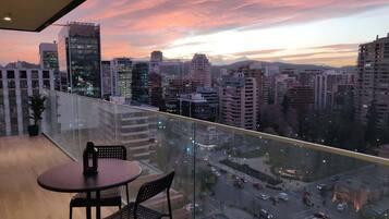 Un balcón con vista a la ciudad al atardecer, con edificios modernos y barandilla de vidrio.