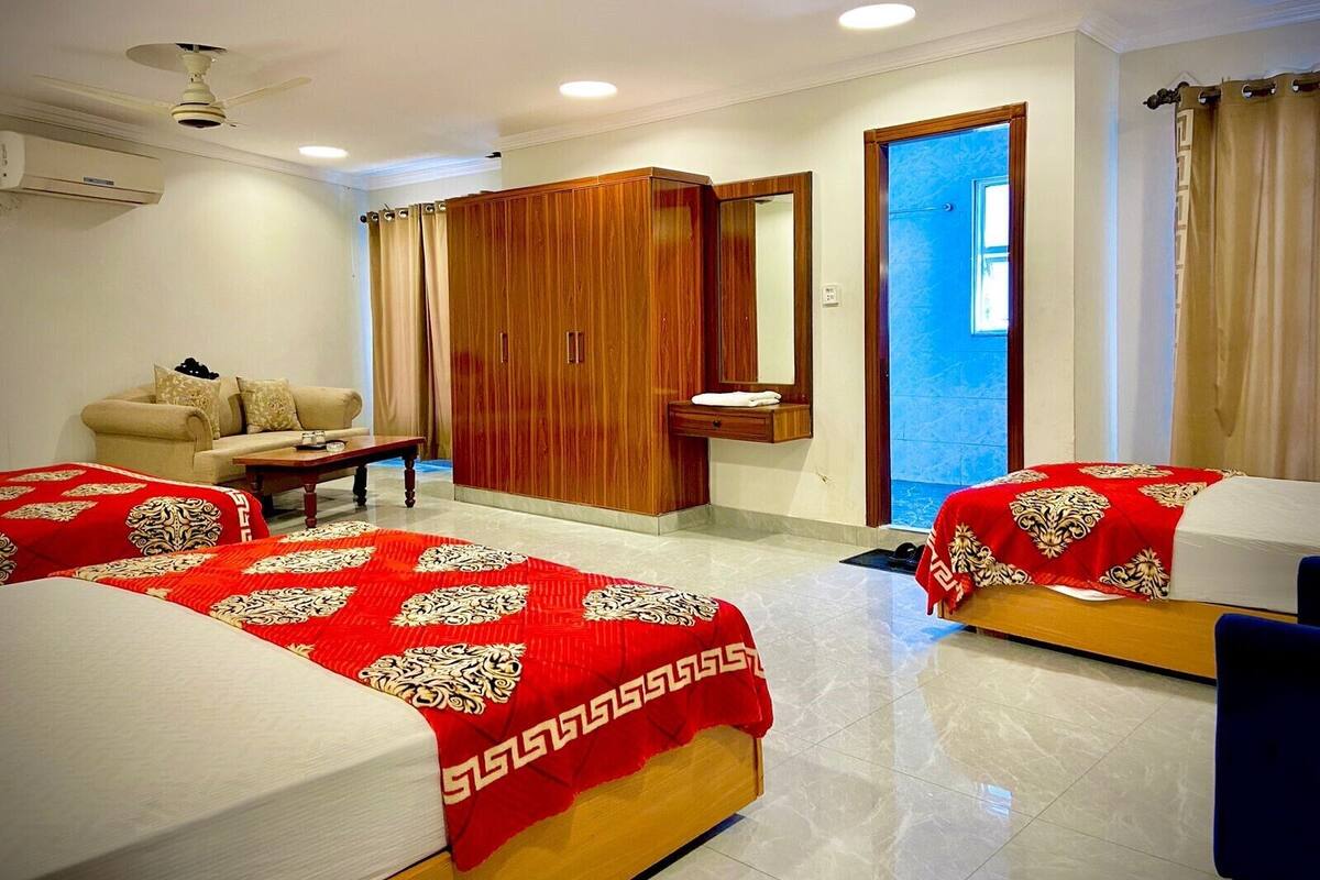 Une chambre d’hôtel avec deux lits, un canapé, une armoire en bois, un miroir et un ventilateur de plafond.