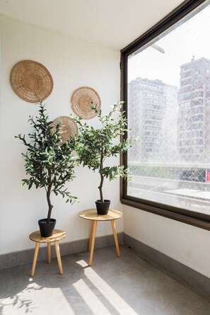 A room with two potted plants on wooden stools, a large window with a view of buildings, and woven wall decorations.