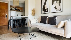 A living room with a sofa, a coffee table, a kitchen area with stools, and abstract art on the wall.