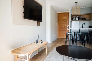 A modern living space with a flat-screen TV mounted on the wall, a wooden bench, a black table, and a kitchen area with stools.