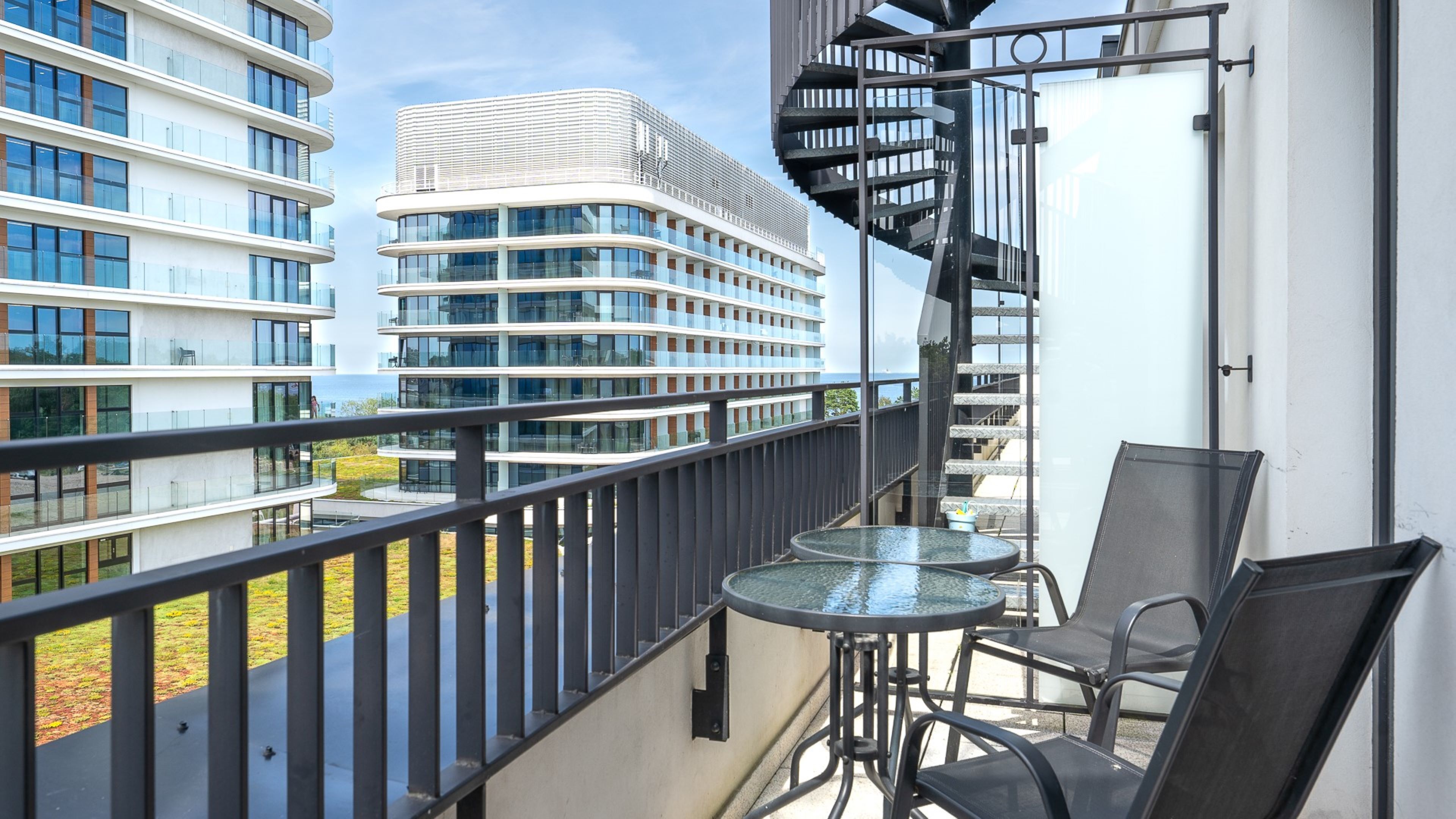 Un balcón con una mesa y sillas de vidrio, con vistas a edificios modernos y una masa de agua.