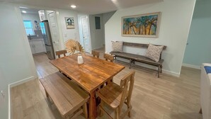 Un comedor con una mesa de madera, bancos y un banco con cojines. Hay un cuadro en la pared y una puerta que da a otra habitación.