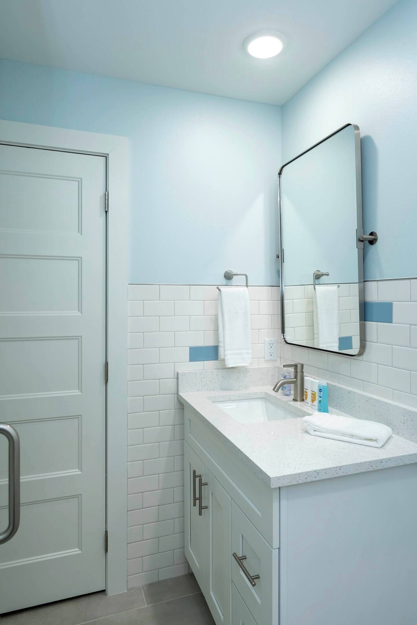 Un baño con paredes de azulejos blancos, un lavabo blanco, un espejo y una puerta.