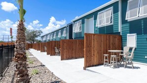 Fila de habitaciones de motel con paneles de madera para privacidad, una palmera y asientos al aire libre.