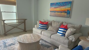 Una sala de estar con un sofá beige, cojines estampados, una mesa de centro redonda y un cuadro con un atardecer sobre un puerto deportivo.