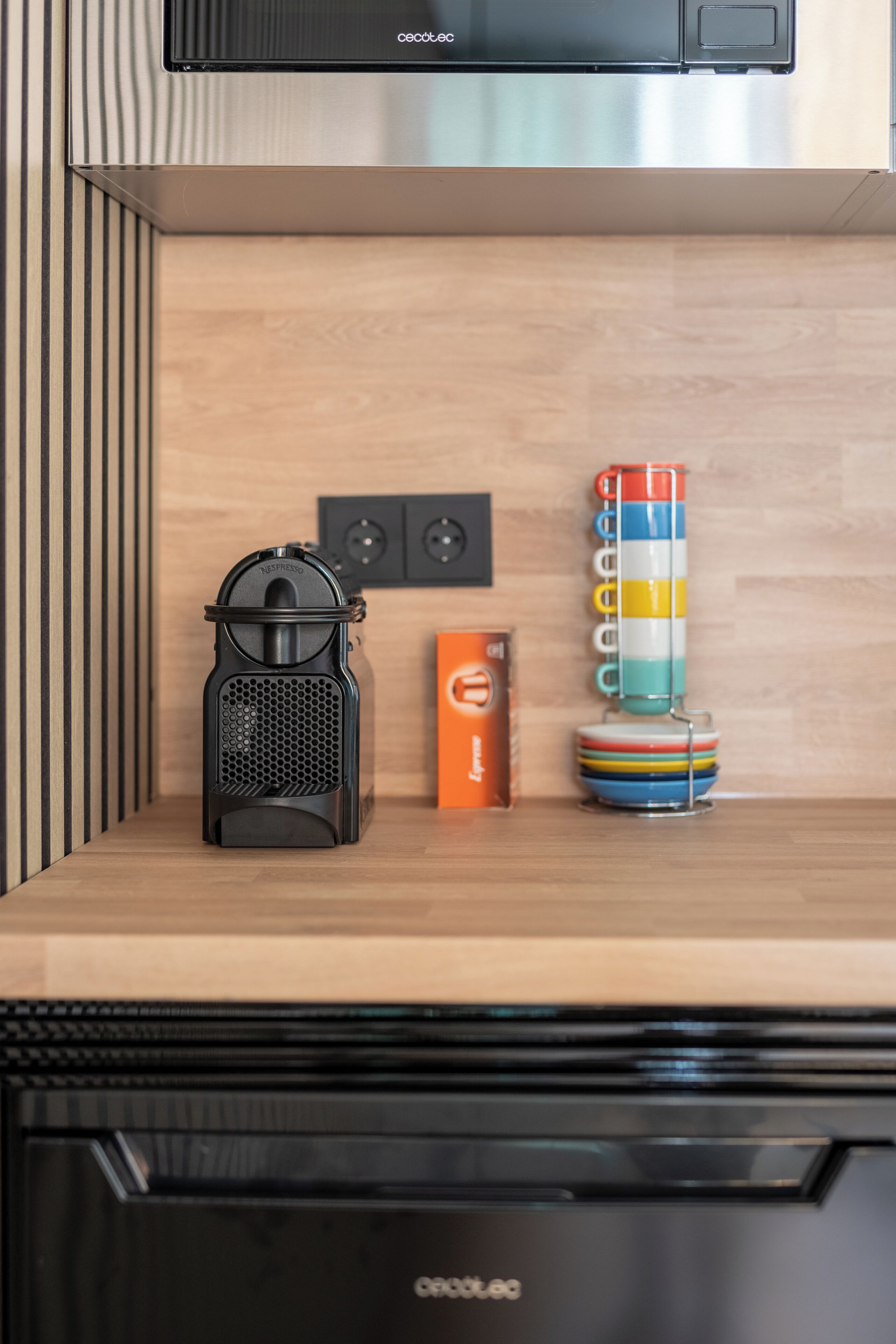 Une cuisine équipée d’une machine à café, d’une pile de tasses colorées et d’une boîte de capsules de café.
