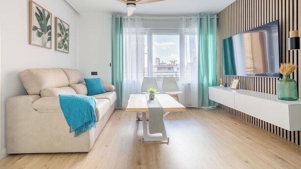 A modern living room with a sofa, a table, a ceiling fan, a TV, and a view of the city.