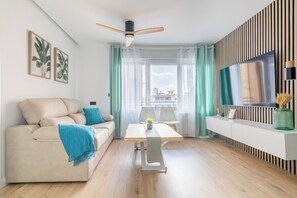 A modern living room with a sofa, a table, a ceiling fan, a TV, and a view of the city. - Luxor Apartment in Parque Sur Hospital - University Area (Albacete)