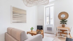 A modern living room with a sofa, a coffee table, a TV, a dining table with chairs, a decorative mirror, and a large window.