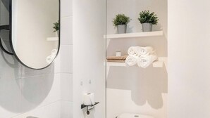A modern bathroom with a white sink, mirror, toilet, and shelves with towels and plants.