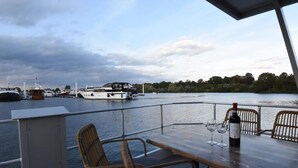 Terraza con sillas de mimbre y una mesa, con vista a un río tranquilo con barcos.