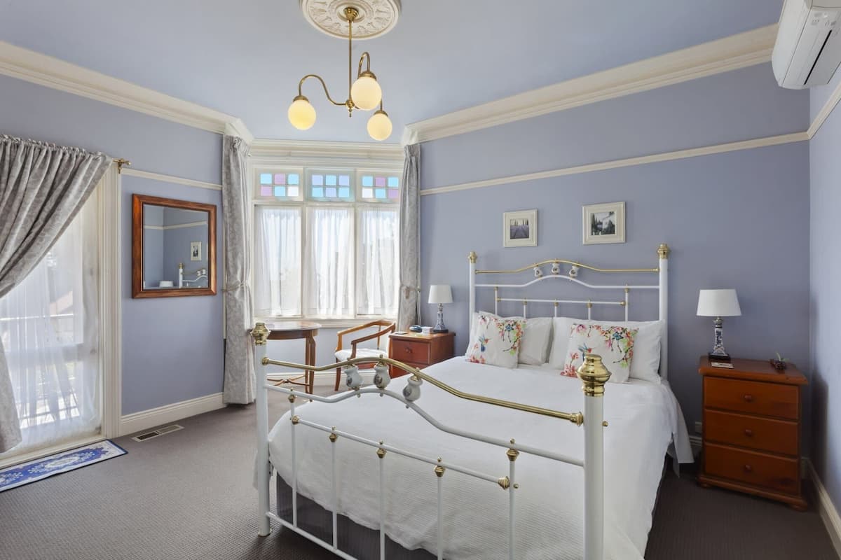 Une chambre à coucher avec un lit blanc, une table de chevet en bois, une commode, un bureau et une chaise. Il y a une fenêtre avec des rideaux, un miroir et un lustre.