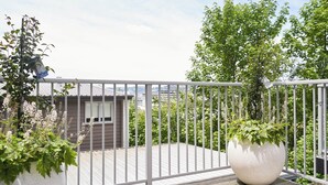 A balcony with a white railing, a large white planter with greenery, and a view of a residential area with trees and buildings.