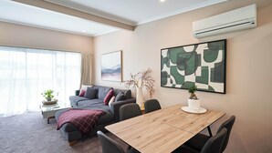 A modern living room with a sofa, a dining table, and chairs. There is a large window with curtains, a potted plant, and an air conditioning unit on the wall.