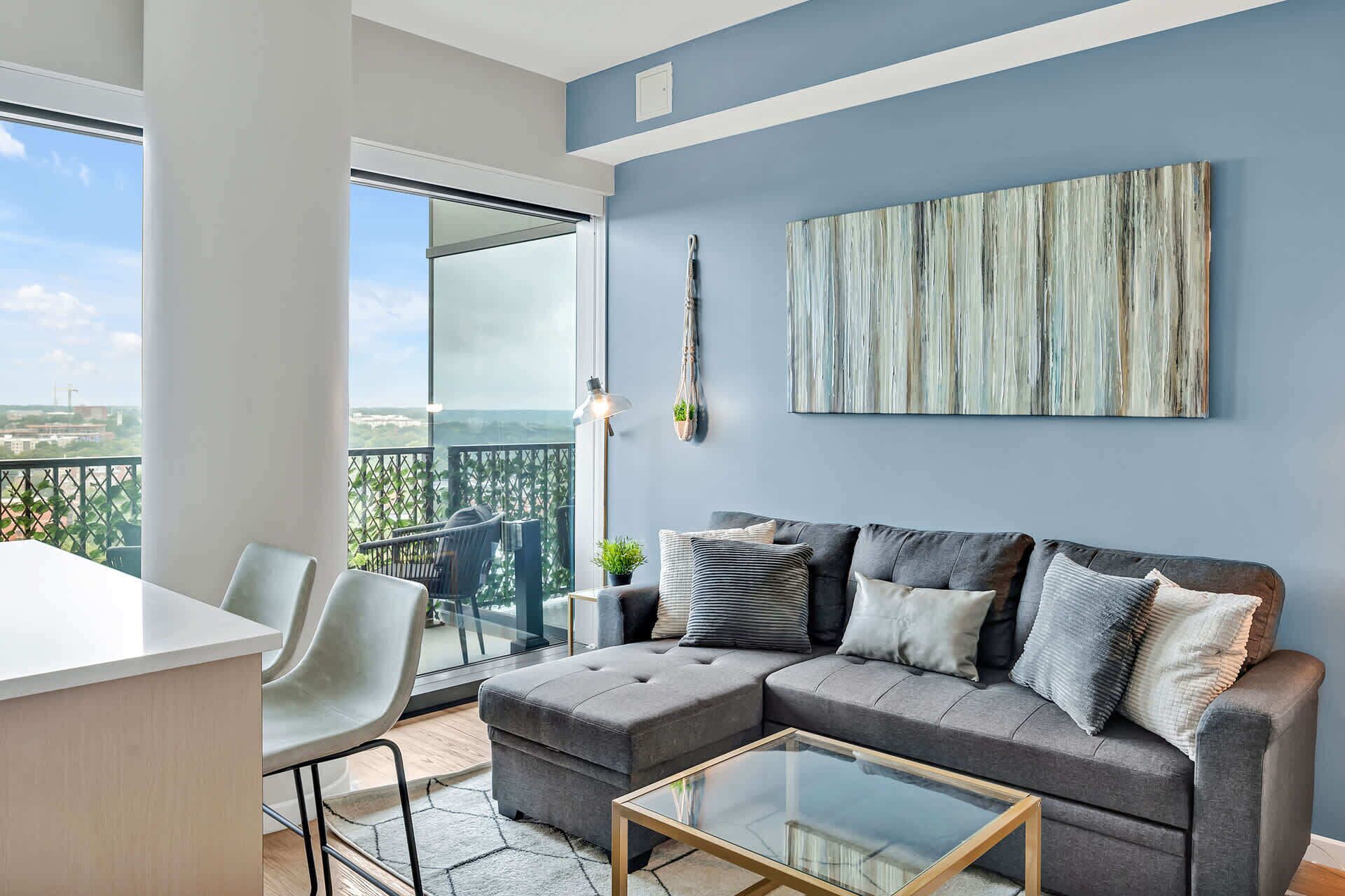 Uma sala de estar moderna com um sofá seccional cinza, uma mesa de centro de vidro e vista para a cidade através de grandes janelas.