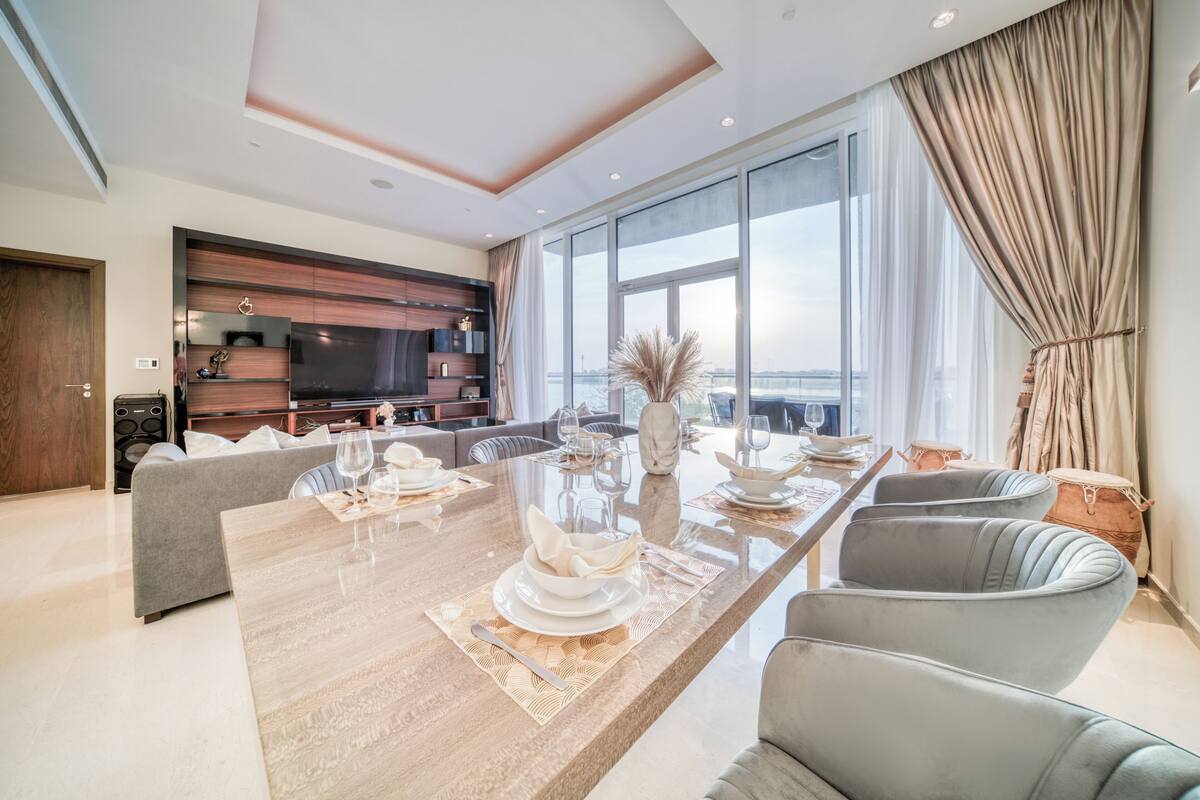 A modern living room with a large dining table set for six, a sofa, and a TV mounted on the wall.