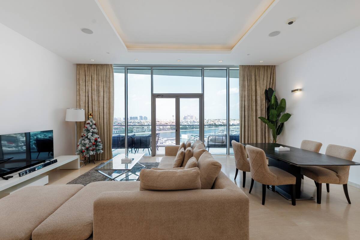 A modern living room with a large flat-screen TV, a beige sofa, a dining table with chairs, and a view of a cityscape through large windows.