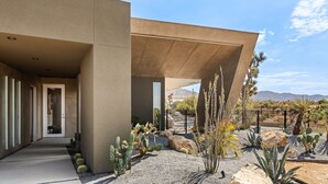 Un bâtiment moderne doté d’une façade en béton, d’une allée couverte et d’un jardin désertique agrémenté de cactus et de rochers.