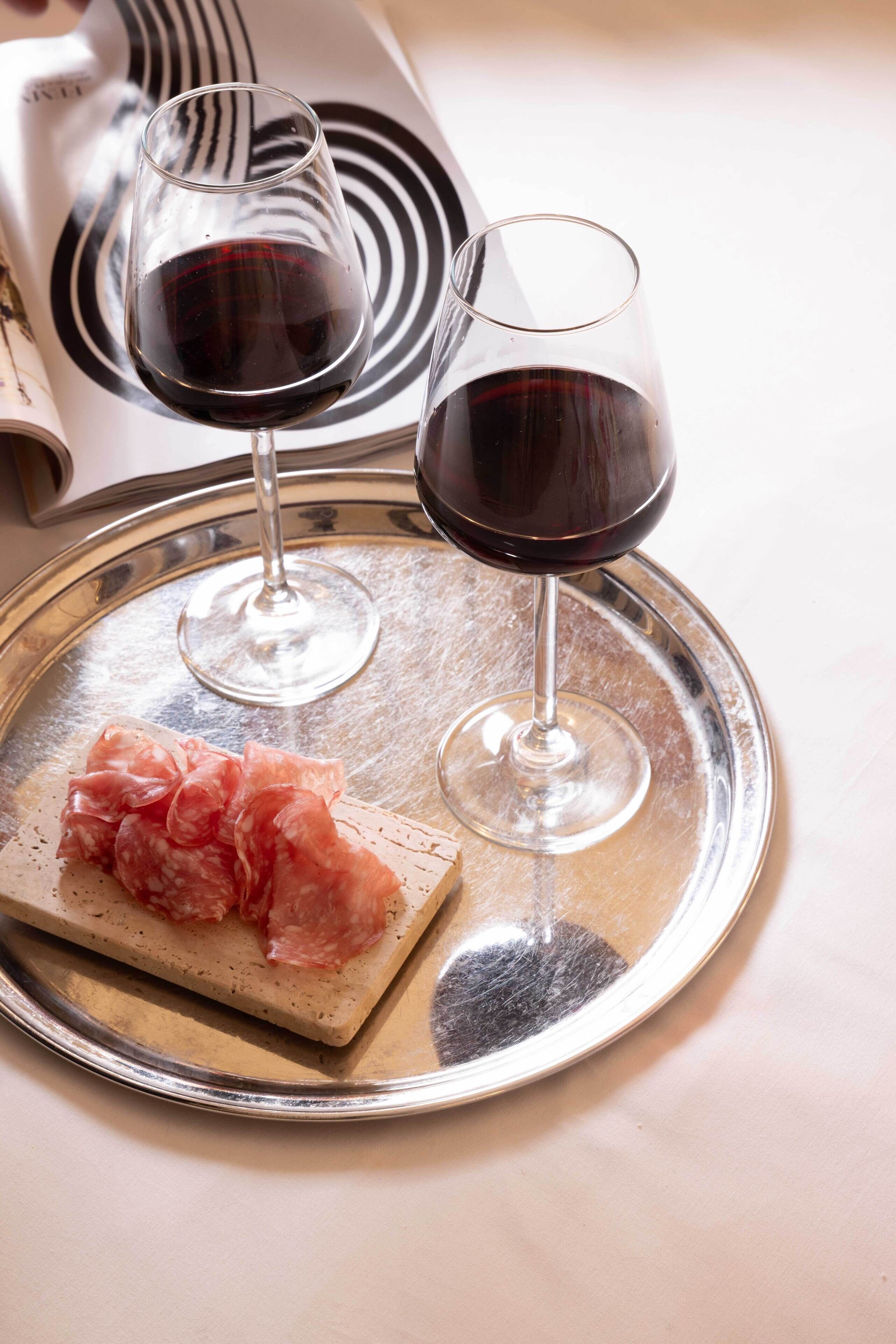 Una bandeja con dos copas de vino tinto y una loncha de embutido sobre una mesa.