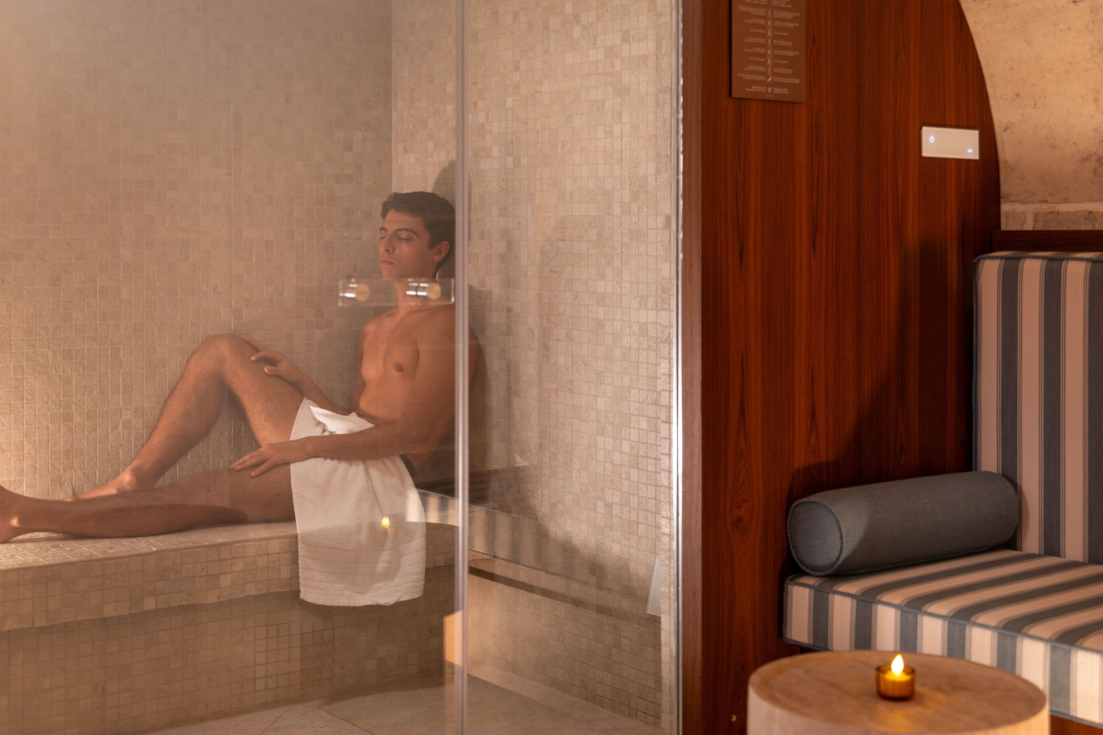 A person relaxing in a glass-enclosed bathtub with a towel, a wooden bench, and a lit candle on a small table.