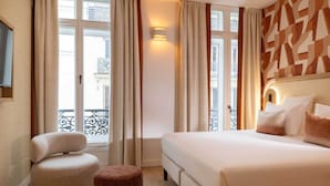 A hotel room with a large bed, a round chair, a small ottoman, and a view of the city through large windows. - Madame Drouot & Spa - Opéra (Paris)
