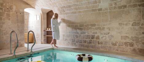 A stone-walled indoor swimming pool with clear water, a metal ladder, and two inflatable rings.