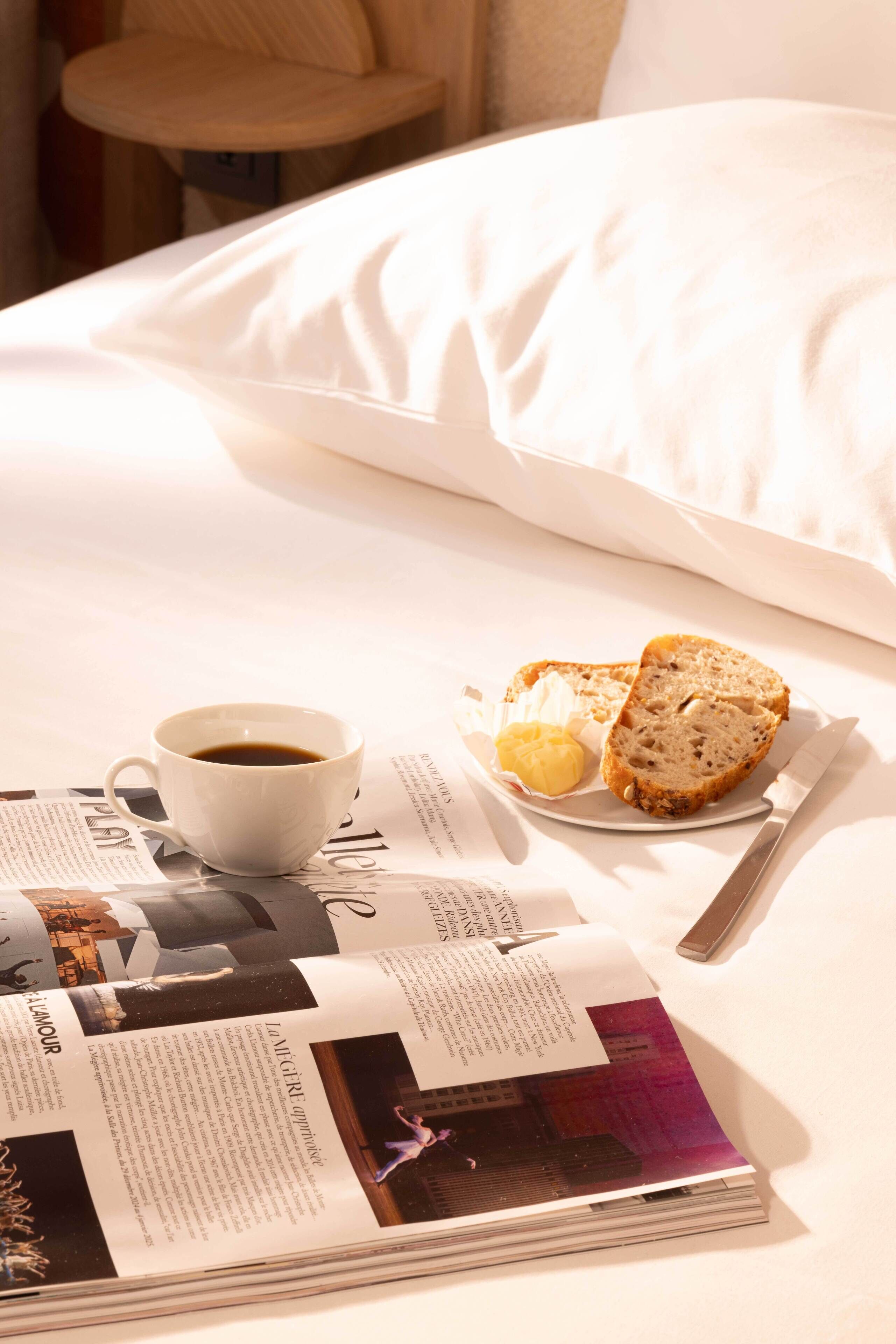Una cama bien tendida con sábanas blancas, una taza de café, un plato con pan y mantequilla, y una revista sobre la cama.