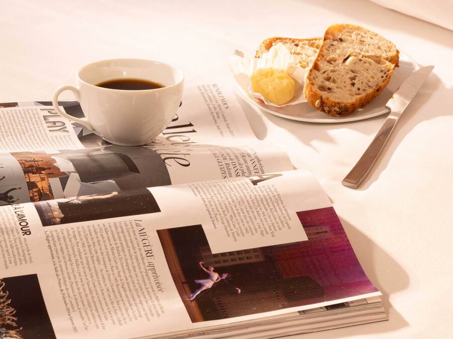 Una cama bien tendida con sábanas blancas, una taza de café, un plato con pan y mantequilla, y una revista sobre la cama.