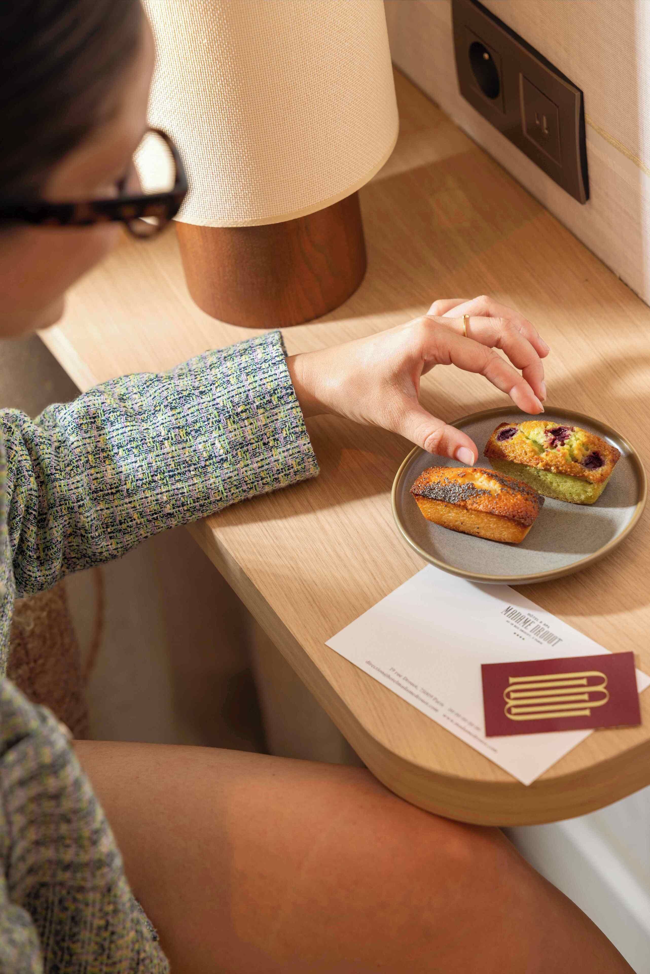 Une personne assise à une table en bois, devant une assiette de muffins et une lampe.
