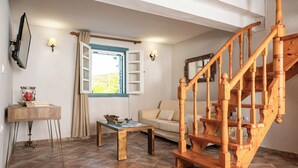 A living room with a wooden staircase, a sofa, a coffee table, and a window with curtains. - THALIA'S COTTAGE - Cozy, Romantic Retreat for Two (Samos)