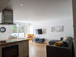 Un salon moderne comprenant un coin cuisine, un téléviseur à écran plat, un canapé et un fauteuil agrémenté d’un coussin orné du drapeau britannique (Union Jack).