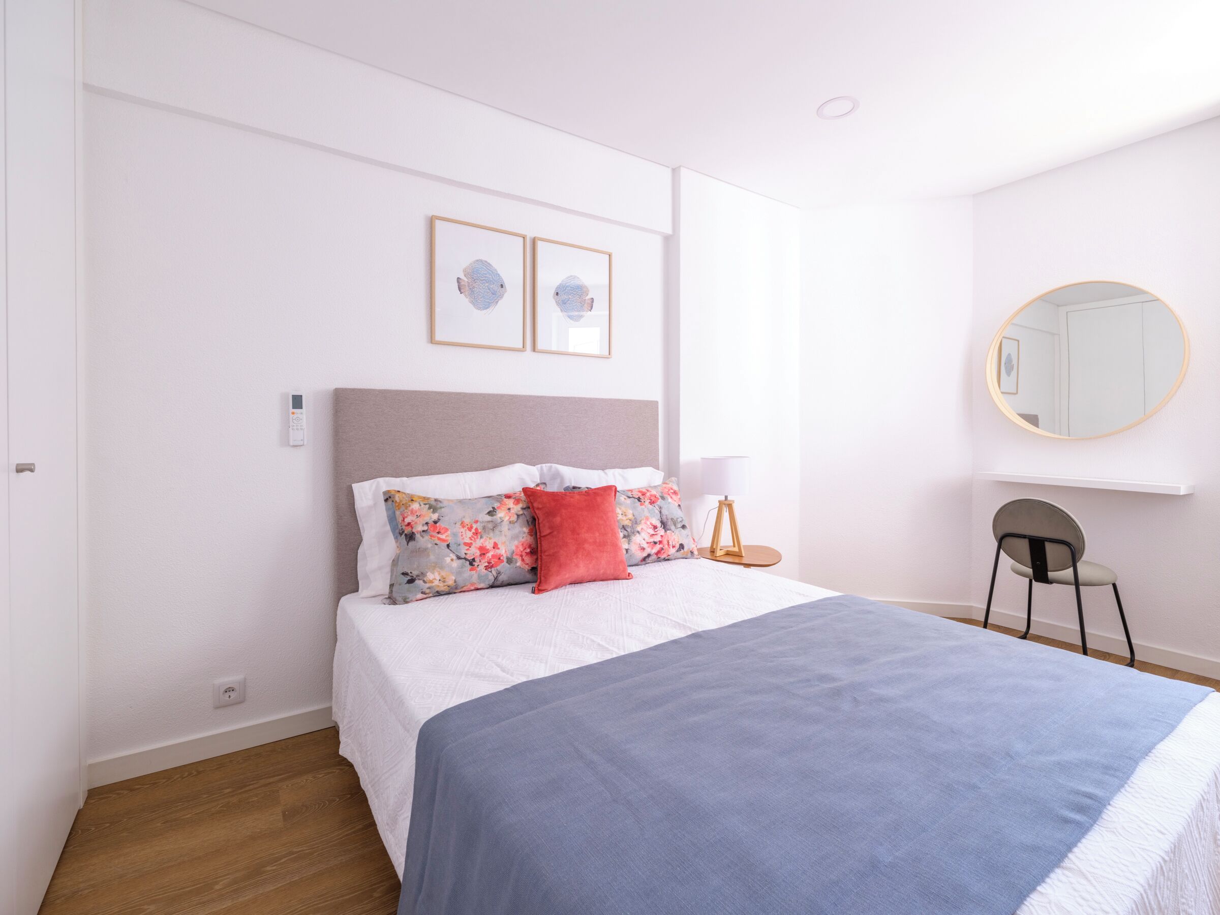 A neatly arranged bedroom with a bed, a bedside table, a lamp, a chair, and a round mirror on the wall.